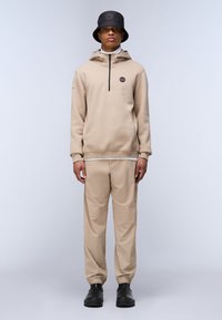 Sudadera beige con media cremallera y logo, combinada con pantalones beige a juego. Un sombrero tipo bucket negro y calzado negro completan el conjunto.