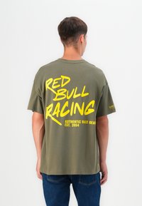 Alyvuogių spalvos medvilninis marškinėliai su dideliu geltonu tekstu ant nugaros: "RED BULL RACING AUTHENTIC RACE GEAR EST. 2004." Atsipalaidavęs siluetas.