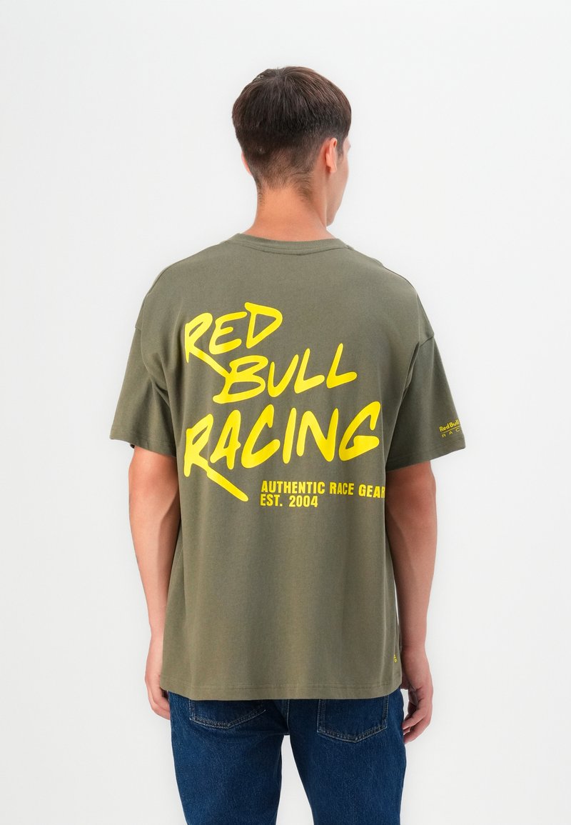 Alyvuogių spalvos medvilninis marškinėliai su dideliu geltonu tekstu ant nugaros: "RED BULL RACING AUTHENTIC RACE GEAR EST. 2004." Atsipalaidavęs siluetas.