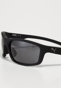 Occhiali da sole rettangolari neri con lenti scure; finitura opaca; presenta un logo metallico Puma sul lato; montatura resistente e design elegante.