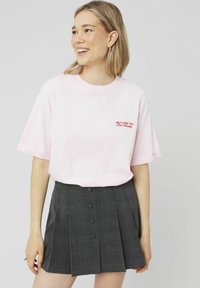 T-shirt en coton rose avec des manches retroussées, arborant un texte rouge sur la poitrine, associé à une jupe plissée grise à carreaux avec des détails de boutons.
