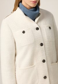 Abrigo blanco texturizado con dos bolsillos de parche, botones negros y un suave cuello de tortuga azul acanalado por debajo. Diseño simple y limpio.
