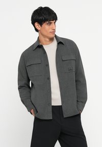 Sötétszürke gombos ing, texturált anyagból készült, két mellzsebbel és címkével. Egy világos szürke pulóver fölött viselve, hétköznapiasan styled.