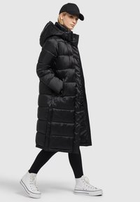 Sort pufferjakke med hætte, der har en kviltet design, lynlås foran og sidelommer, båret over sorte leggings og hvide sneakers.
