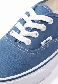 Sininen Vans-tenniskenkä valkoisilla nauhoilla, valkoinen kumipohja, näkyvät омpeleet ja pieni Vans-logo-tagi sivulla.