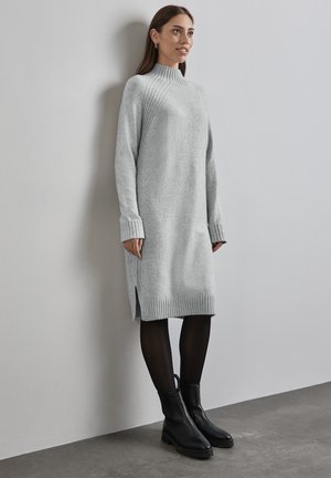 Helles graues Strickkleid mit hohem Kragen, gerippten Bündchen und seitlichen Schlitzen. Tragen mit schwarzen Stiefeletten und schwarzen Strumpfhosen. Glatte Textur.