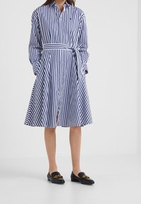 Kvinna som bär en blå och vit randig skjortklänning med en knuten midja och svarta loafers med guldkedjedetaljer.