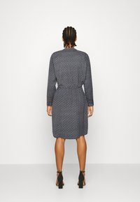 Vestido azul oscuro con estampado geométrico, mangas largas, cintura ajustada y dobladillo a la altura de la rodilla. Llevado con zapatos de tacón negros.