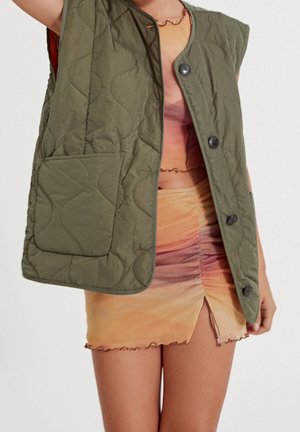Veste matelassée sans manches vert olive avec de gros boutons et une poche, portée sur une mini-jupe froncée multicolore et un top court.