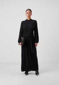 Črna rebrasta dolgoročna obleka s prilegajočim pasom, dolžina do tal, v kombinaciji z majhno črno torbico in gležnjarji.