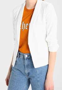 Witte blazer met een getailleerd ontwerp, open voorkant en lange mouwen, gecombineerd met een oranje grafisch T-shirt en een spijkerbroek met hoge taille.