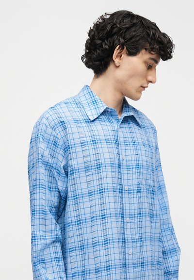 Νέος άνδρας με σγουρά σκούρα μαλλιά, ντυμένος με ανοιχτό μπλε καρό πουκάμισο μακρυμάνικο, γυρισμένος πλευρικά μπροστά σε ένα απλό φόντο.