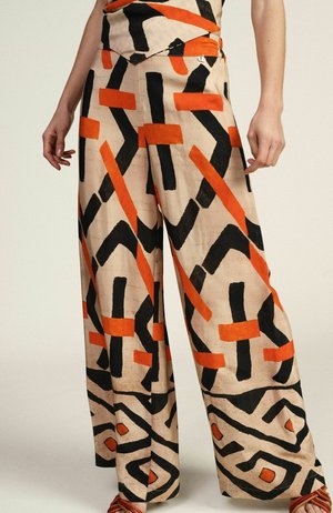 Pantaloni beige a gamba larga con motivi geometrici astratti neri e arancioni audaci, indossati con top abbinato e sandali marroni.