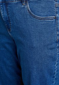 Jean en denim de teinte bleu foncé, présentant une coupe classique, une poche avant et des coutures contrastées le long des coutures. La texture semble lisse.