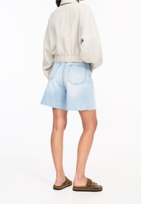 Veste en lin gris clair coupée, portée sur un short en denim bleu clair aux bords effilochés, associée à des sandales marron à enfiler ornées de clous.