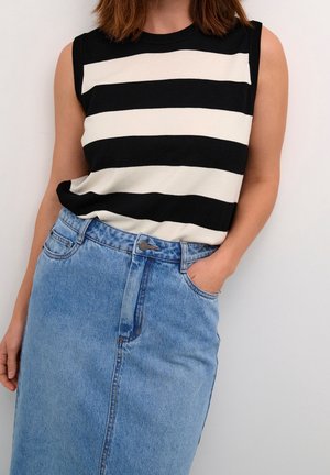 Kvinde i ærmeløs sort-hvidstribet top med blå denimnederdel, den ene hånd i lommen, stående mod en ensfarvet lys baggrund.