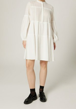 Vestido blanco hasta la rodilla hecho de tela ligera con pliegues, abertura con botones y mangas largas abullonadas. Se lleva con mocasines negros y calcetas.