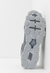 Sohle eines grauen Nike-Sneakers mit kreisförmigen Profilmustern, kreuzförmigen Rillen und dem Nike-Logo auf einem weißen Hintergrund.