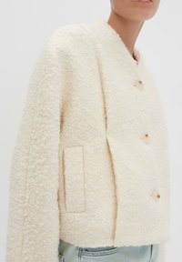 Vrouw in een crèmekleurige, gestructureerde wollen jas met grote knopen en zijzakken over lichtblauwe jeans.