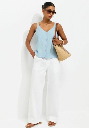 Mujer con top sin mangas con botones de color azul claro, pantalones anchos blancos, sandalias marrones, gafas de sol y lleva un bolso tejido marrón.