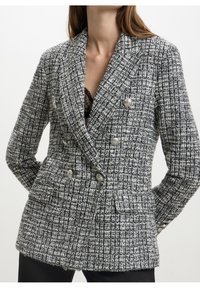 Femme portant un blazer croisé noir et blanc texturé avec des boutons argentés et un haut en dentelle noire en dessous.