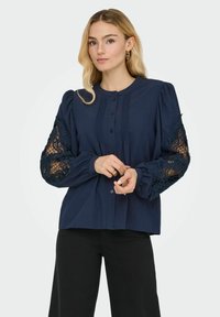 Blusa blu navy con bottoni, maniche a sbuffo, con dettagli in pizzo trasparente sulle maniche e pieghe verticali sul davanti. Tessuto testurizzato.