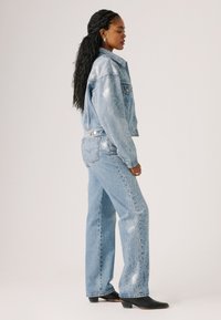 Lichte blauwe denim jasje en spijkerbroek set. Het jasje heeft subtiele versieringen; de spijkerbroek heeft een sierlijk ontwerp langs de zijkant. Zwarte enkellaarsjes.