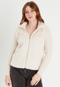 Chaqueta de felpa sintética con cremallera en color crema y mangas de punto acanalado. Incluye bolsillos laterales y un suave cuello, creando un contraste texturizado en el diseño.