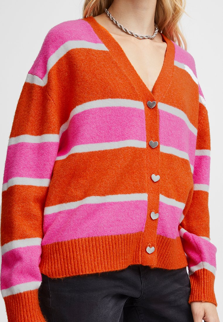 Kobieta nosząca kardigan z dekoltem w kształcie litery V w szerokie pomarańczowe, różowe i białe paski, z guzikami w kształcie serca, zestawiony z czarnymi spodniami.