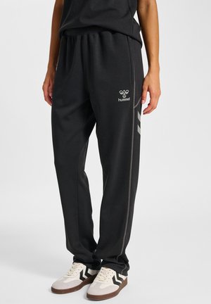 Osoba nosząca czarne dresy Hummel z bocznymi pasami oraz beżowe sneakersy z czarnymi wzorami w jodełkę na jednolitym tle.