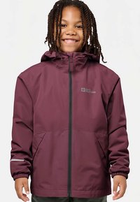 Enfant souriant avec des cheveux tressés portant une veste à capuche Jack Wolfskin de couleur bordeaux avec fermeture éclair sur le devant et poignets élastiques.