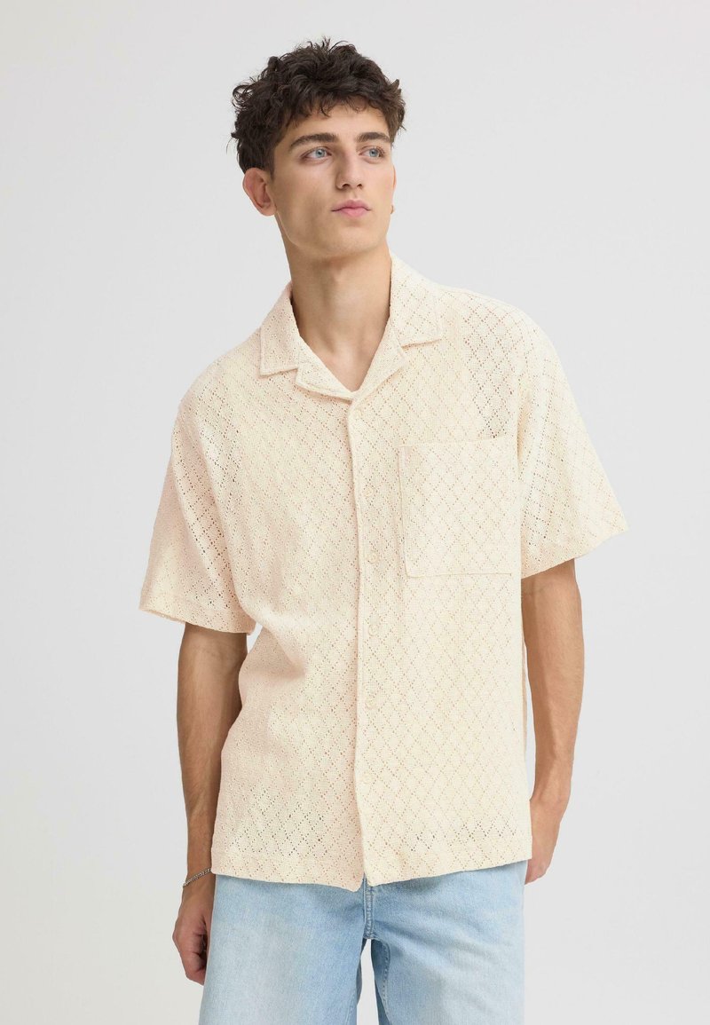 Chemise à manches courtes couleur pêche clair, texturée avec un motif en losange, col ouvert et poche poitrine, associée à un jean bleu clair.