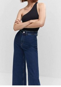 Bodysuit negro de un solo hombro combinado con jeans de talle alto y pierna ancha en azul oscuro. Los vaqueros cuentan con un cierre de botón y cremallera metálicos clásicos.