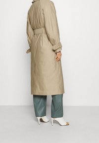 Trench coat beige con cintura, maniche lunghe, spacco laterale e polsini abbottonati, abbinato a pantaloni a gamba larga color teal e mule bianche.
