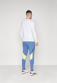 Long-sleeve white top and blue leggings with yellow accents and side patterns, paired with white and black cleats.