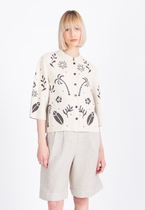 Vrouw met een blonde boblijn draagt een crèmekleurig jasje met zwarte botanische patronen en lichtbeige knielange shorts.