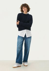 Navy knitted sweater with ribbed neckline and cuffs, layered over a white shirt; paired with wide-leg blue jeans and white sneakers.