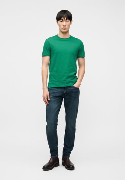 Νεαρός άνδρας όρθιος, φορώντας πράσινο κοντομάνικο μπλουζάκι, σκούρα μπλε τζιν και καφέ δερμάτινα μποτάκια, μπροστά σε απλό ανοιχτό φόντο.