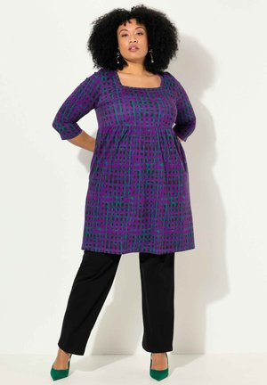 Vestido con estampado púrpura y verde, de mangas tres cuartos y escote cuadrado, combinado con pantalones negros y tacones verdes.