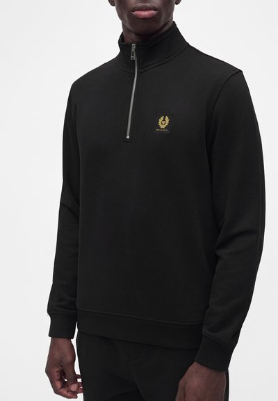 Homme portant un pull noir à manches longues avec une fermeture éclair sur le devant et un petit emblème doré sur la poitrine gauche.