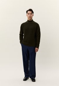 Fiatal férfi áll, sötétzöld strukturált pulóvert, sötétkék nadrágot és fekete elegáns cipőt visel semleges háttér előtt.