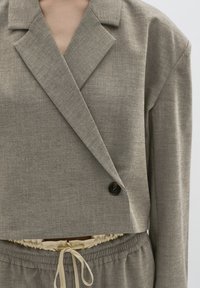 Šedý blazer s krátkym strihom, jemnou textúrou a zapínaním na dva gombíky. V páre s rovnako šedými teplákmi so sťahovacou šnúrkou v uvoľnenom strihu.