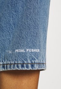 Ourlet de short en jean bleu avec texte blanc brodé "PEDAL PUSHER" au-dessus de la peau visible.