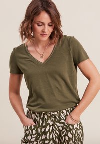 T-shirt verde oliva a maniche corte con scollo a V, caratterizzato da cuciture a contrasto. Combinato con pantaloni larghi a fantasia in verde e bianco.