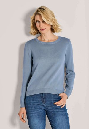 Blonde vrouw met kort haar in een lichtblauwe lange mouwen trui en blauwe spijkerbroek, staande tegen een effen lichte achtergrond.
