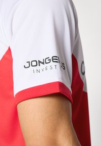 Weißes Sporttrikot mit roten Akzenten. Hat eine glatte Textur, gerippten Stoff an den Ärmeln und das aufgedruckte Logo "JONGEELS INVEST."