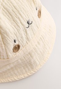 Bombažna klobučasta kapa svetlo rumene barve z teksturirano tkanino. Na njej je vezen obraz medveda z rjavim nosom in črnimi brki.