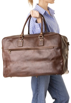 Donna che indossa una camicia a righe blu, che tiene una grande borsa da viaggio in pelle marrone per i manici al suo fianco.