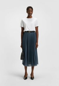Witte t-shirt, marineblauwe geplooide midi-rok, zwarte riem en zwarte pumps met spitse neus. Model houdt een kleine zwarte handtas vast. Eenvoudig ontwerp.