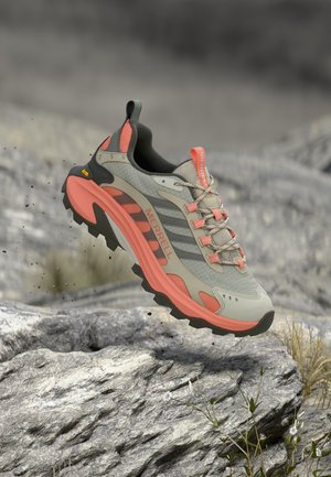 Chaussure de trail avec des accents gris, noirs et corail flottant au-dessus d'un terrain rocheux avec de petites plantes et des fleurs.